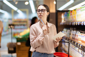 Portrait of young thoughtful caucasian woman wearing eyeglasses holds product list and look around....