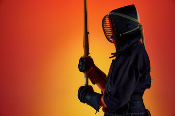 Side view. Portrait of man, professional kendo athlete in uniform posing with shinai sword against gradient studio background in neon light. Concept of martial arts, sport, Japanese culture, motion