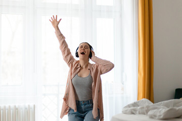 Young Caucasian happy woman wearing headphones dance and singing in living room. Window in background. Home weekend