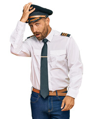 Handsome man with beard wearing airplane pilot uniform surprised with hand on head for mistake,...
