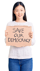 Young beautiful chinese woman holding save our democracy cardboard banner looking positive and happy standing and smiling with a confident smile showing teeth