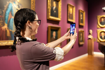 Young Caucasian woman takes selfie or makes video call with smartphone. Visiting gallery or museum....