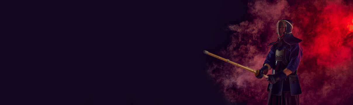 Man, professional kendo athlete, combat sportsman posing with shinai sword against gradient dark background in neon light with smoke. Concept of martial arts, sport, Japanese culture. Banner
