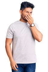 Handsome latin american young man wearing casual tshirt smelling something stinky and disgusting,...