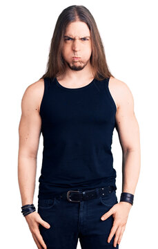 Young adult man with long hair wearing goth style with black clothes puffing cheeks with funny face. mouth inflated with air, crazy expression.