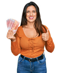 Beautiful hispanic woman holding norwegian krone banknotes smiling happy and positive, thumb up doing excellent and approval sign