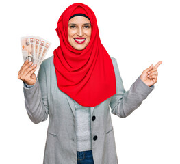 Beautiful hispanic woman wearing islamic hijab holding 10 pounds banknotes smiling happy pointing with hand and finger to the side