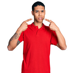 Young hispanic man pointing mouth with fingers clueless and confused expression. doubt concept.