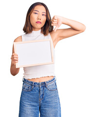 Young beautiful chinese girl holding empty white chalkboard with angry face, negative sign showing dislike with thumbs down, rejection concept