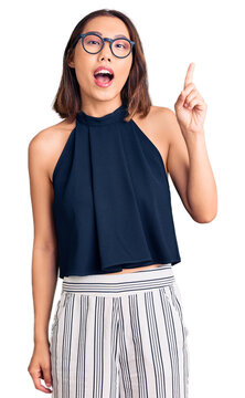 Young beautiful chinese girl wearing casual clothes and glasses pointing finger up with successful idea. exited and happy. number one.