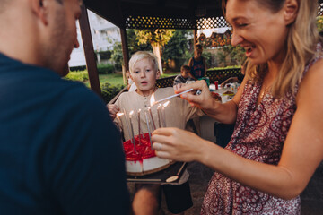 Parents holding Birthday cake for their 11 year old son, woman is lighting candles on cheesecake with red jelly