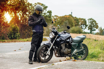 Fototapeta motorcyclist in a helmet and motorcycle clothing stands at an old cafe racer with a smartphone obraz