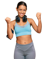 Beautiful hispanic woman wearing sportswear and headphones screaming proud, celebrating victory and success very excited with raised arms
