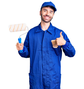 Young hispanic man wearing painter uniform holding roller smiling happy and positive, thumb up doing excellent and approval sign