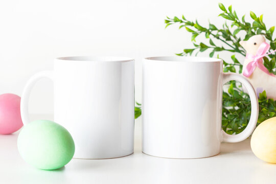 Easter mockup of two white mugs on table with colorful painted eggs and greenery. Blank coffee cups mock up.