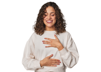 Radiant young woman with stunning curls laughs happily and has fun keeping hands on stomach.