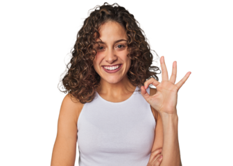 Radiant young woman with stunning curls winks an eye and holds an okay gesture with hand.