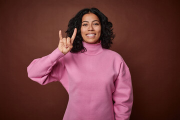 Young colombian curly hair woman isolated on brown background showing a horns gesture as a revolution concept.