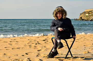 A little boy with coat and hat sitting on chair on the beach
