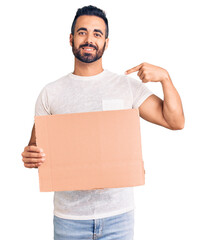 Young hispanic man holding banner cardboard pointing finger to one self smiling happy and proud