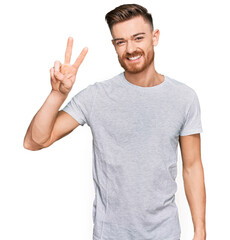 Young redhead man wearing casual grey t shirt showing and pointing up with fingers number two while smiling confident and happy.