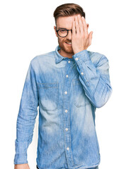 Young redhead man wearing casual denim shirt covering one eye with hand, confident smile on face and surprise emotion.