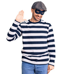 Young handsome man wearing burglar mask waiving saying hello happy and smiling, friendly welcome gesture