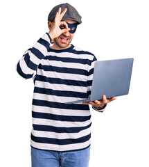 Young handsome man wearing burglar mask using laptop smiling happy doing ok sign with hand on eye looking through fingers