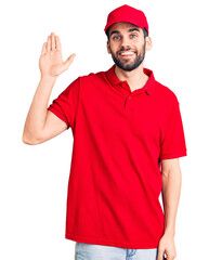 Young handsome man with beard wearing delivery uniform waiving saying hello happy and smiling, friendly welcome gesture