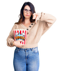 Beautiful young brunette woman eating popcorn with angry face, negative sign showing dislike with thumbs down, rejection concept