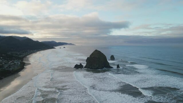 Wide bird eye drone aerial view on Haystack Rock in Cannon Beach, Oregon, United States. Dark rock formation in water. Cloudy dramatic weather. Waves splashing. High quality 4k footage