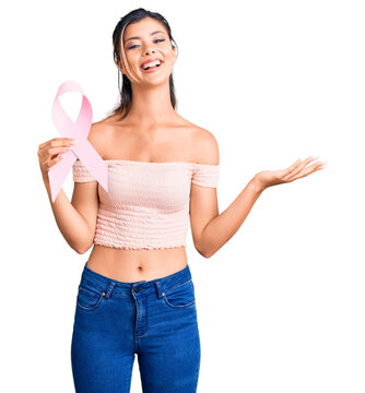 Young beautiful woman holding cancer awareness pink ribbon celebrating victory with happy smile and winner expression with raised hands