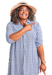 Young african american plus size woman wearing summer hat cheerful with a smile of face pointing with hand and finger up to the side with happy and natural expression on face