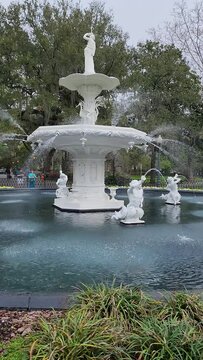 panning footage of a white stone water fountain surrounded by yellow and white flowers and lush green trees and plants with people walking at Forsyth Park in Savannah Georgia USA