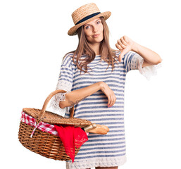 Beautiful caucasian woman wearing summer hat and holding picnic wicker basket with bread with angry face, negative sign showing dislike with thumbs down, rejection concept