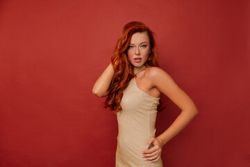Pleased caucasian lady wears golden dress is posing in studio. Indoor portrait of cheerful red-haired girl with wavy hairstyle isolated on red background.