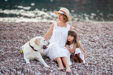 Fototapeta premium Pregnant mother with little daughter are sitting along the sand of the beach near the sea with golden retriever. Millennial woman holds dog on leash, girl by the hand. Concept of traveling with pets