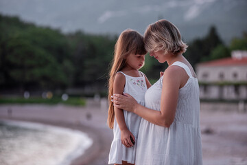 Obraz premium Close-up portrait of pregnant mother with little daughter walking hand in hand in white dresses at sunset by the sea. Child care. Healthy motherhood. The concept of vacation, travel single parent.