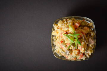 Traditional Soviet salad Olivier served in glass bowl. Salad with potato and carrot cubes, green peas, pickled cucumbers, eggs with green spring onion scales on top. Dark background. Horizontal image