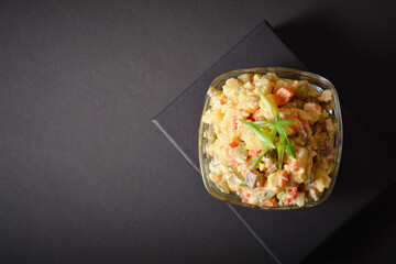 Above of Traditional Russian salad Olivier served in glass bowl located on black box. Salad with potato and carrot cubes, green peas, pickled cucumbers, eggs on Dark background. Dark mood style