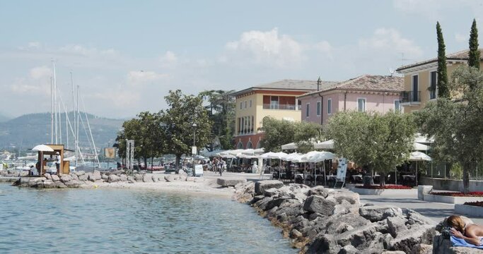 Cisano, Italy - 06 29 2021: Cisano Port on Garda Lake, Bardolino. Harbour of Cisano near Lazise and Verona. Famous Porticciolo with tourists on bicycles and moored banks. Still shot and wide angle.