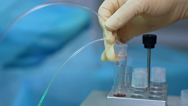 Gloved hand holding a tube connected to the test tube. Liquid is flowing through the tube filling the reservoir. Close up. Blurred backdrop.