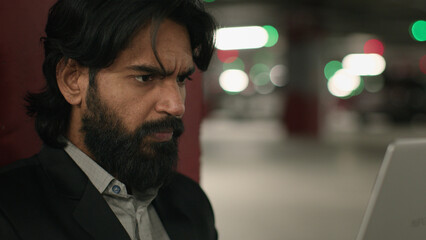 Close-up puzzled serious Indian man looking at laptop screen in underground parking focused worried...