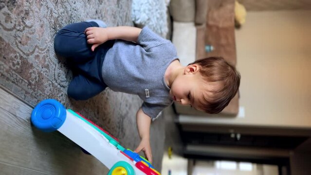 Healthy Caucasian baby boy playing in the room. Adorable kid sits on the floor touching the toy with his finger. Vertical screen.