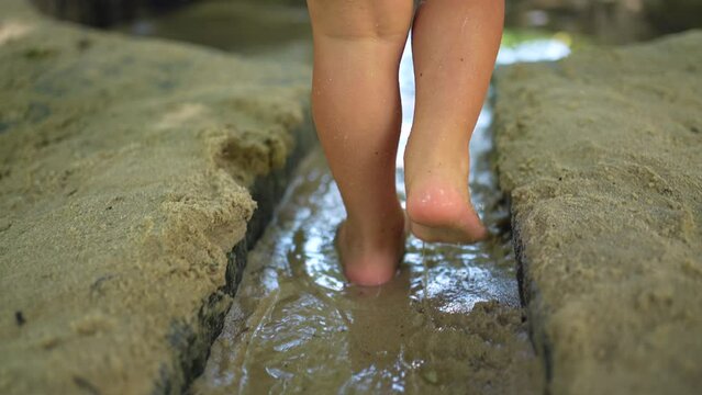 Child walking barefoot outside in water mud. Closeup kid feet stepping in wet sand outdoors and jumping into puddle at park during hot summer day