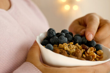 close-up female hand takes berries on blurred background with bokeh, healthy food white cup, oatmeal, blueberries, nuts, almonds, peanuts , raisins, dried fruits, raw food diet, vegetarian food