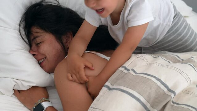 Little boy son trying to wake up mom in morning bed. Child waking up sleeping mother during weekend. Authentic real life family lifestyle