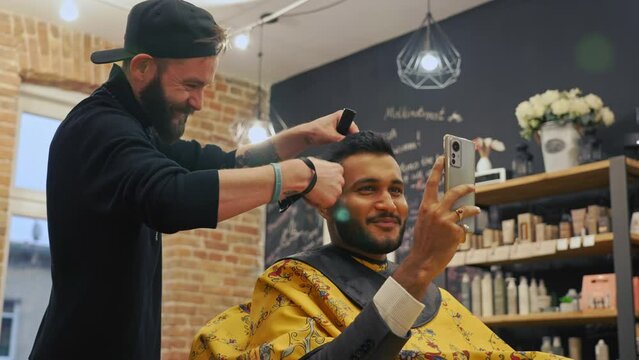 Hair salon's client fliming himself while having his hair cut by a male hair expert. Another hairdresser walking in the background. Horizontal video. High quality 4k footage
