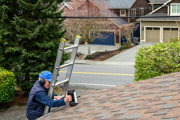 Senior man wearing hearing protection and carrying a canister of moss killer, climbing an aluminum extension ladder up to a rooftop 