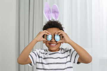 Cute African American boy in bunny ears headband covering eyes with Easter eggs indoors